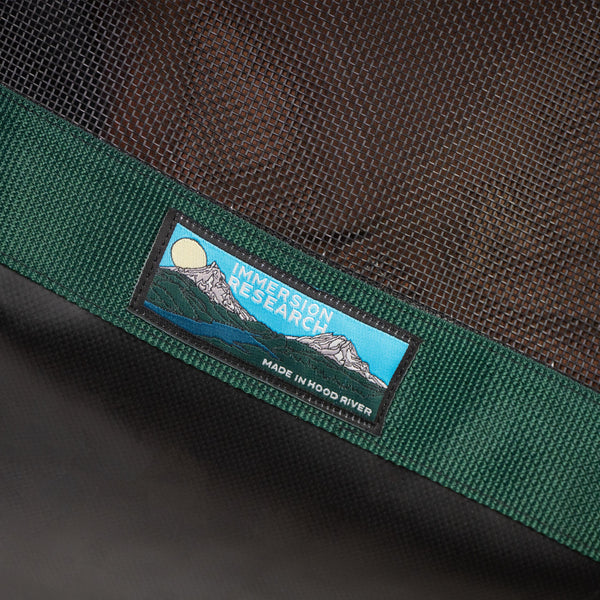 Close-up of a green woven strap with a rectangular embroidered patch reading "IMMERSION RESEARCH" and "MADE IN HOOD RIVER," set against a dark brown mesh background.