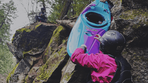 Person in Aphrodite Dry Suit in Cactus Flower holding a blue kayak against a rocky background