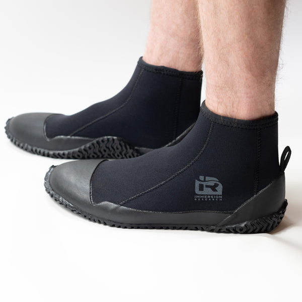 A person wearing black neoprene boot covers with black rubber soles, shown from the calves down on a clean white studio background. A gray IR logo.