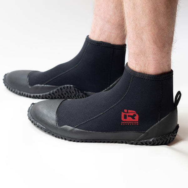 A person wearing black neoprene boot covers with black rubber soles, shown from the calves down on a clean white studio background. A red IR logo.