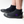 A person wearing black neoprene boot covers with black rubber soles, shown from the calves down on a clean white studio background. A purple IR logo.