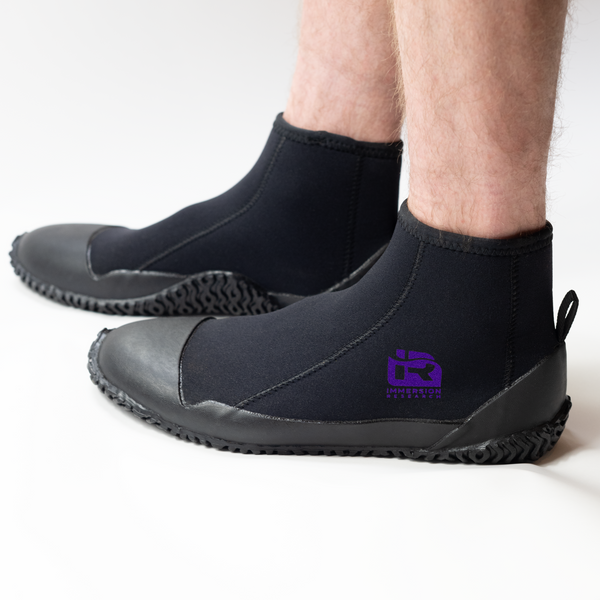 A person wearing black neoprene boot covers with black rubber soles, shown from the calves down on a clean white studio background. A purple IR logo.