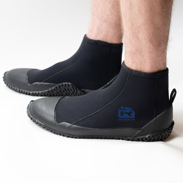 A person wearing black neoprene boot covers with black rubber soles, shown from the calves down on a clean white studio background. A blue IR logo.