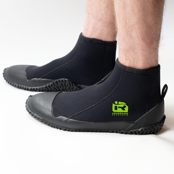A person wearing black neoprene boot covers with black rubber soles, shown from the calves down on a clean white studio background. A green IR logo.