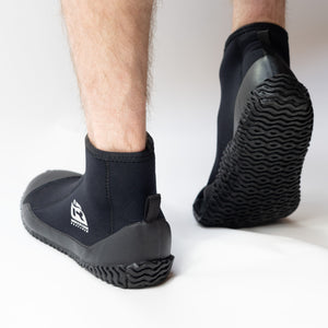 A person wearing black neoprene boot covers with black rubber soles, shown from the calves down on a clean white studio background. A white IR logo.