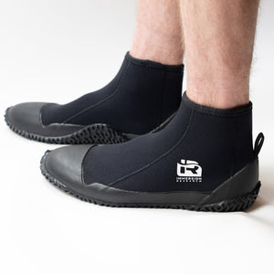 A person wearing black neoprene boot covers with black rubber soles, shown from the calves down on a clean white studio background. A white IR logo.
