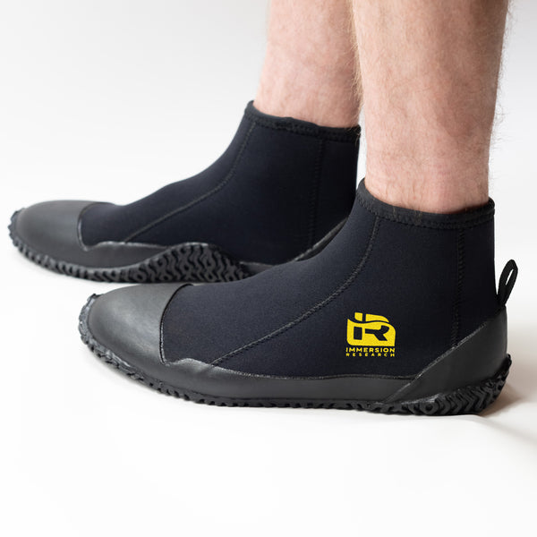 A person wearing black neoprene boot covers with black rubber soles, shown from the calves down on a clean white studio background. A yellow IR logo.