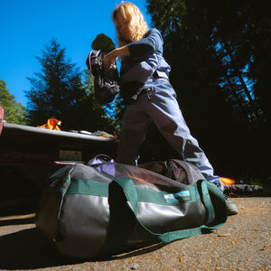 A person packing gear into a large black and green duffel bag, with trees and blue sky in the background.