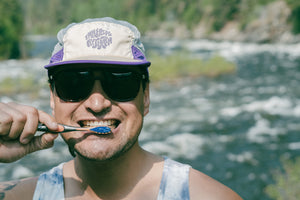 Man brushing his teeth wearing a hat by the river.